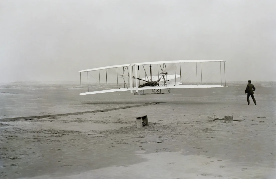 Le Wright Flyer quitte le rail de lancement ; Wilbur court à côté tandis qu’Orville pilote.